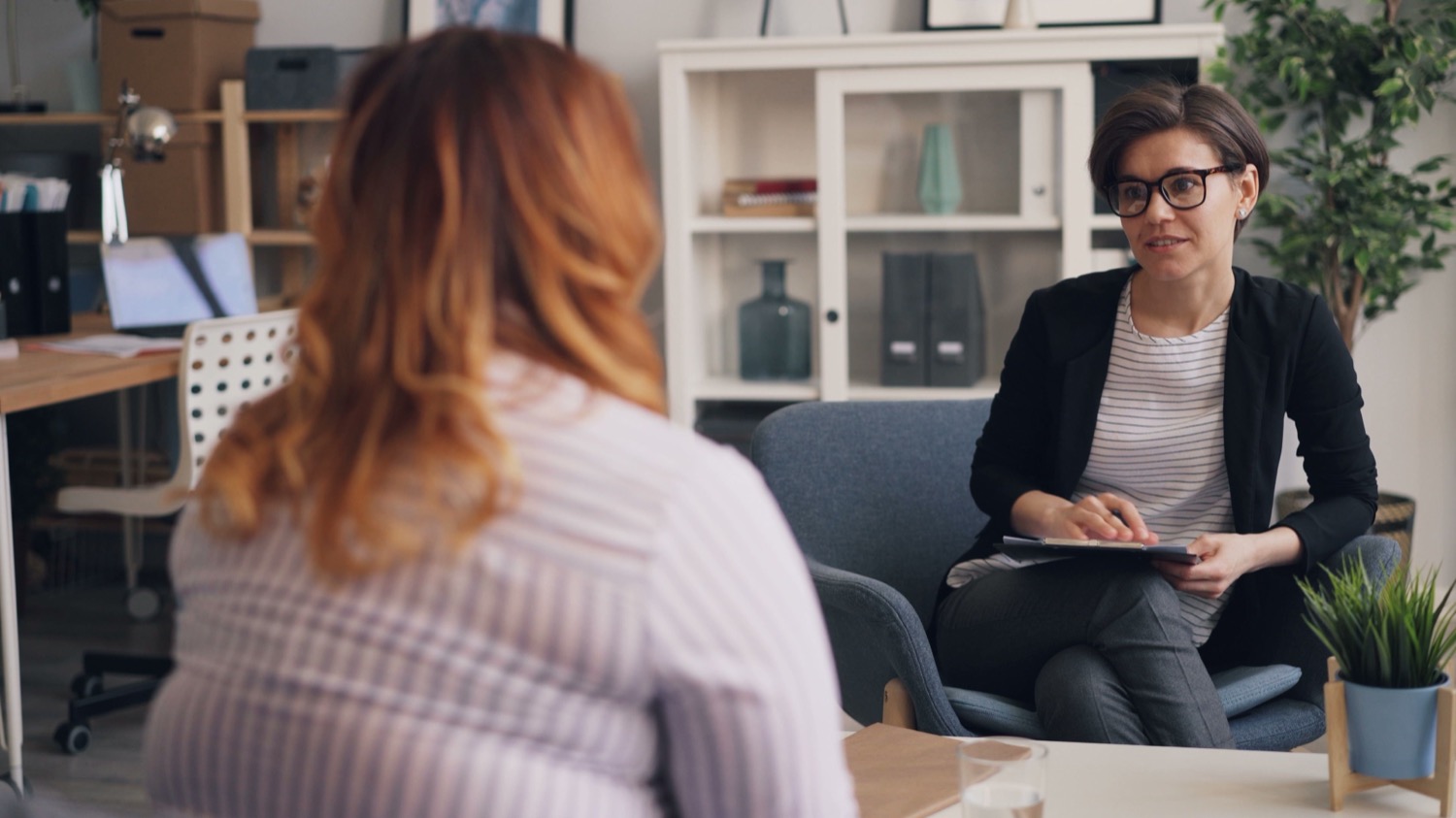 Two women in conversation in an office, with one listening and holding a clipboard.