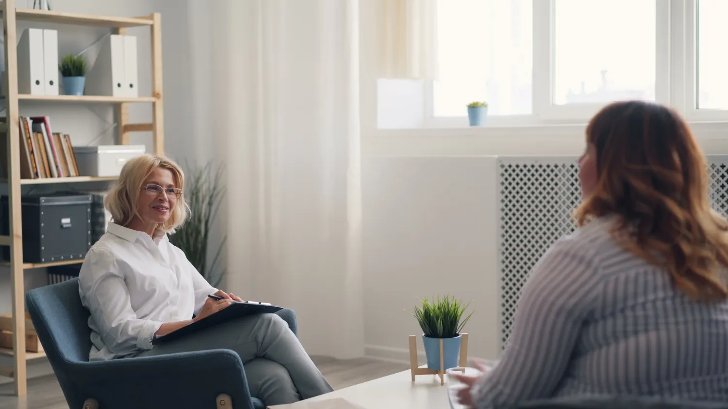 A counselor with a clipboard smiles while speaking with a woman in a sunlit office.